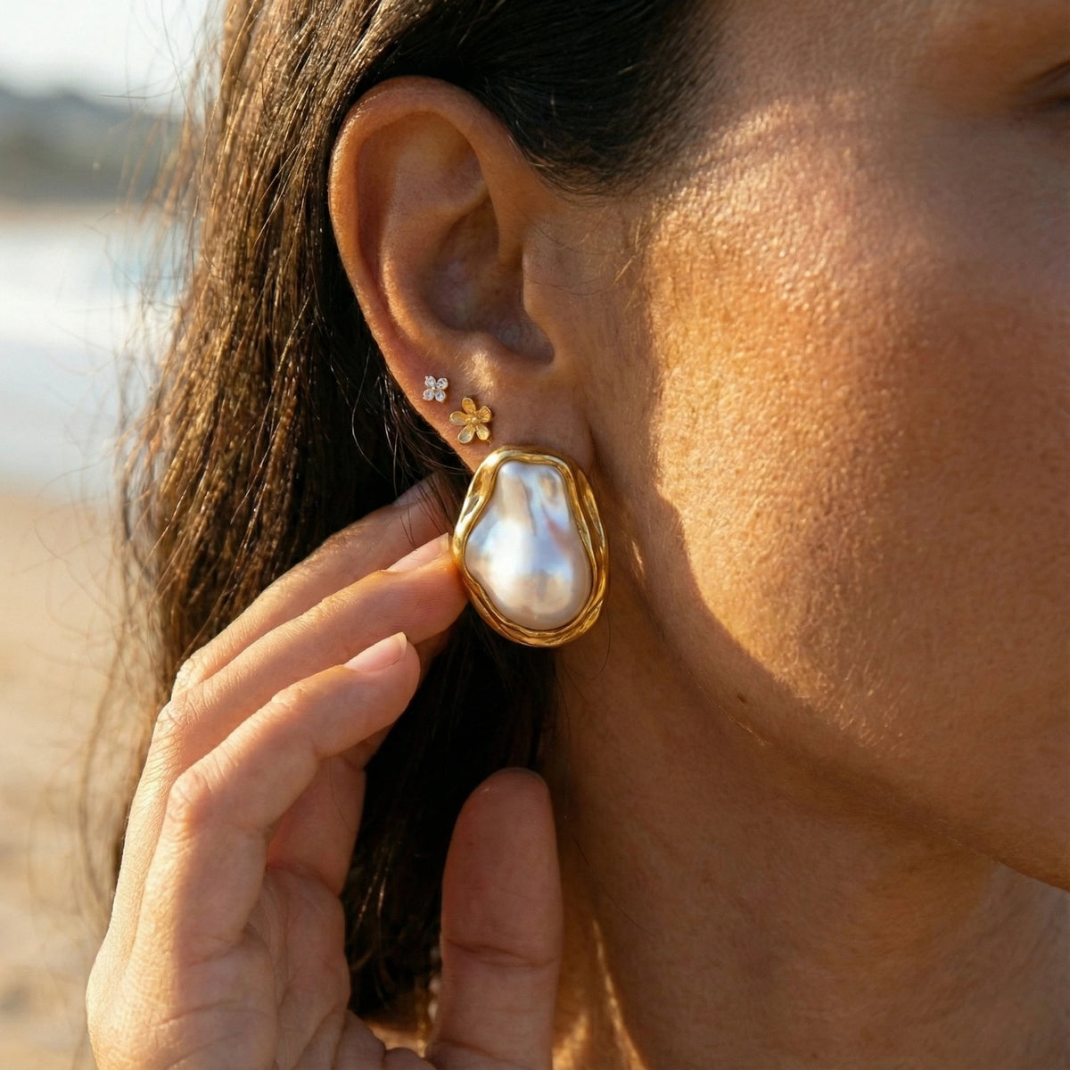 Close-up of a woman wearing a gold Sienne baroque pearl earring on her earlobe, her fingertips resting near her cheek