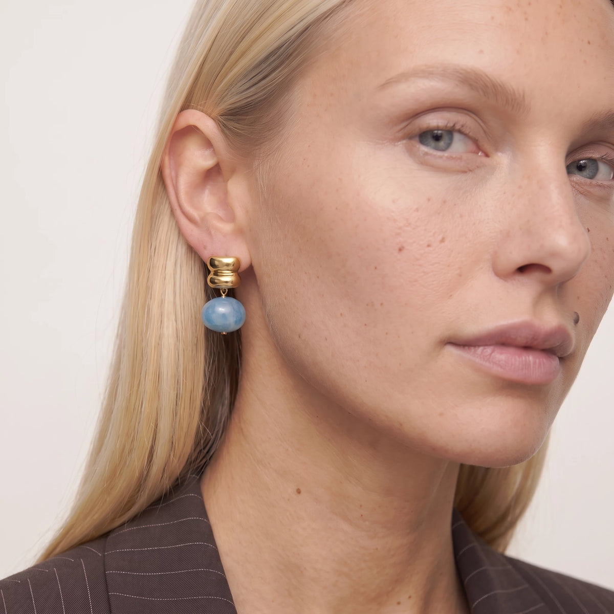 Close-up of a woman with blonde hair wearing an Ondine earring, a blue resin drop under a stacked gold stud, in a pinstripe blazer