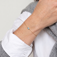 Wrist in a white shirt and grey jumper wearing the Juno bracelet in 14K gold-plated waterproof stainless steel, with a small light blue baguette stone