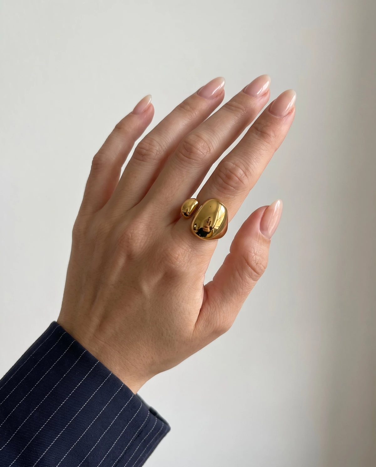 Hand with natural nails wearing the gold Romy dome ring on the middle finger, a navy pinstripe sleeve at the wrist