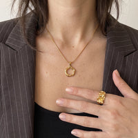 Woman in a brown pinstripe blazer and black top wearing the Runa gold halo pendant necklace, one hand wearing a gold ring