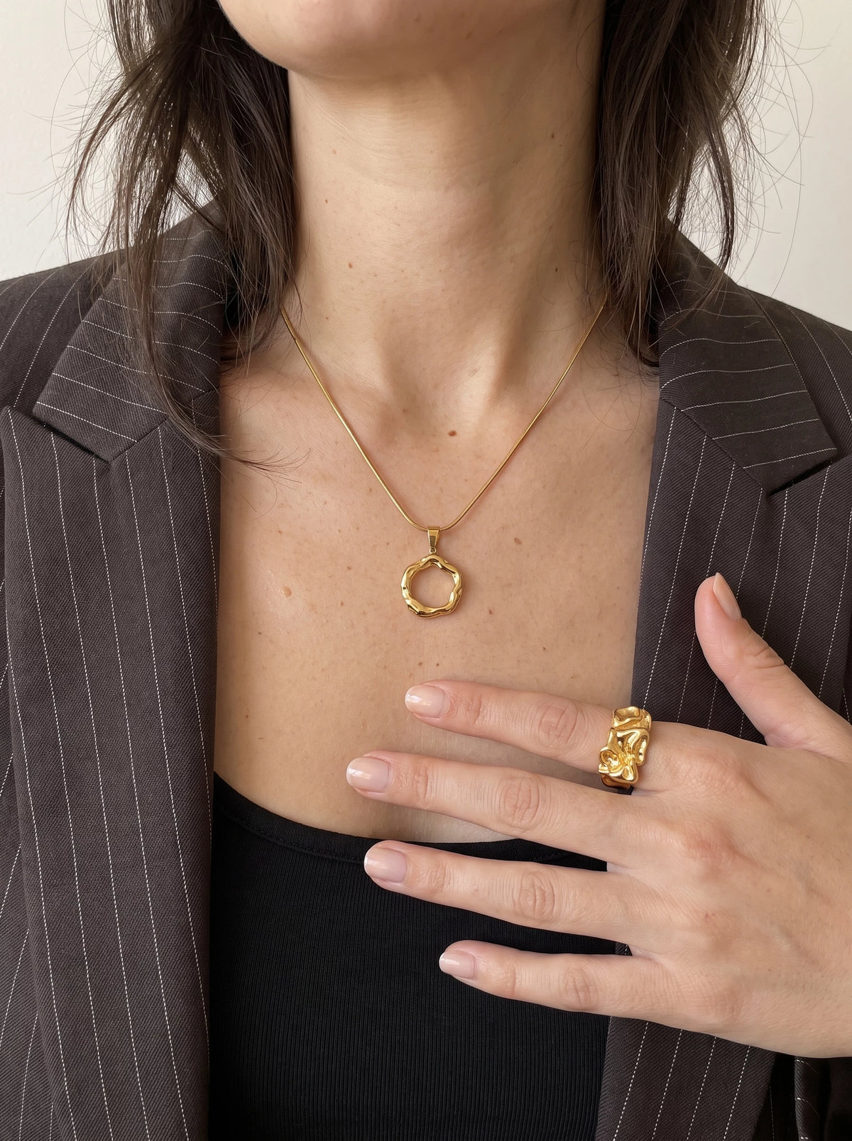 Woman in a brown pinstripe blazer and black top wearing the Runa gold halo pendant necklace, one hand wearing a gold ring