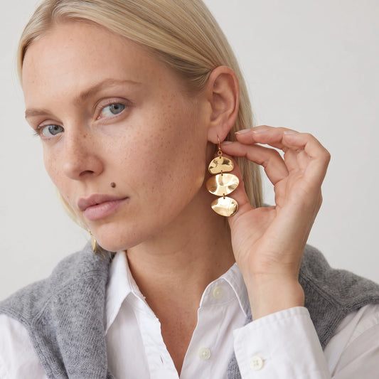 Woman with blonde hair and a beauty mark touching a gold Greta triple disc drop earring, in a white shirt and grey jumper