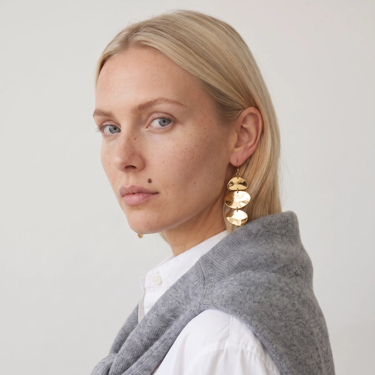 Woman with her hair back wearing the gold Greta triple disc drop earring, a white shirt with a grey jumper over her shoulders