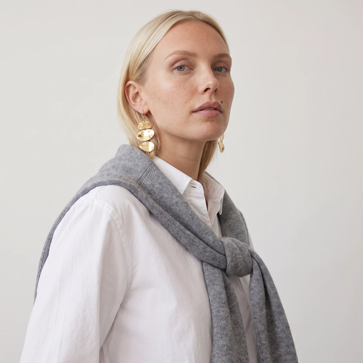 Woman in a white shirt and grey jumper, side view, wearing the gold Greta triple disc drop earring