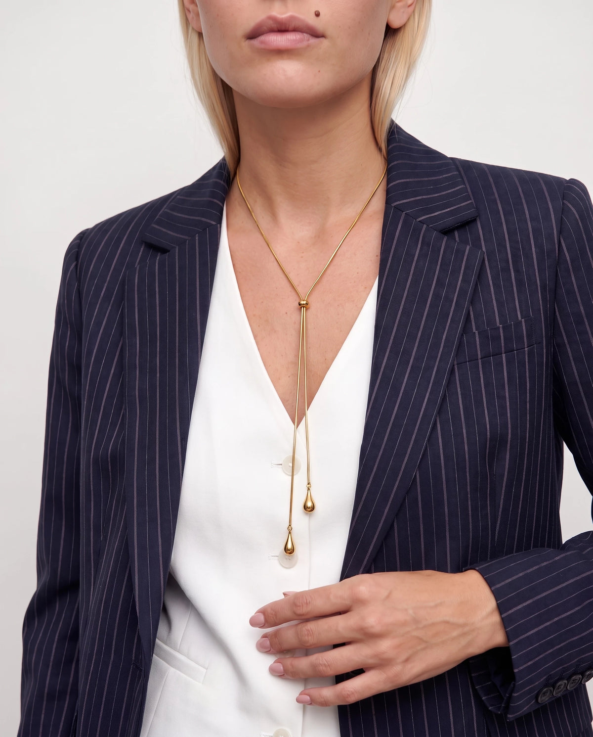 Woman in a navy pinstripe blazer and white vest wearing the gold Elodie lariat, its teardrop drops falling to mid-chest