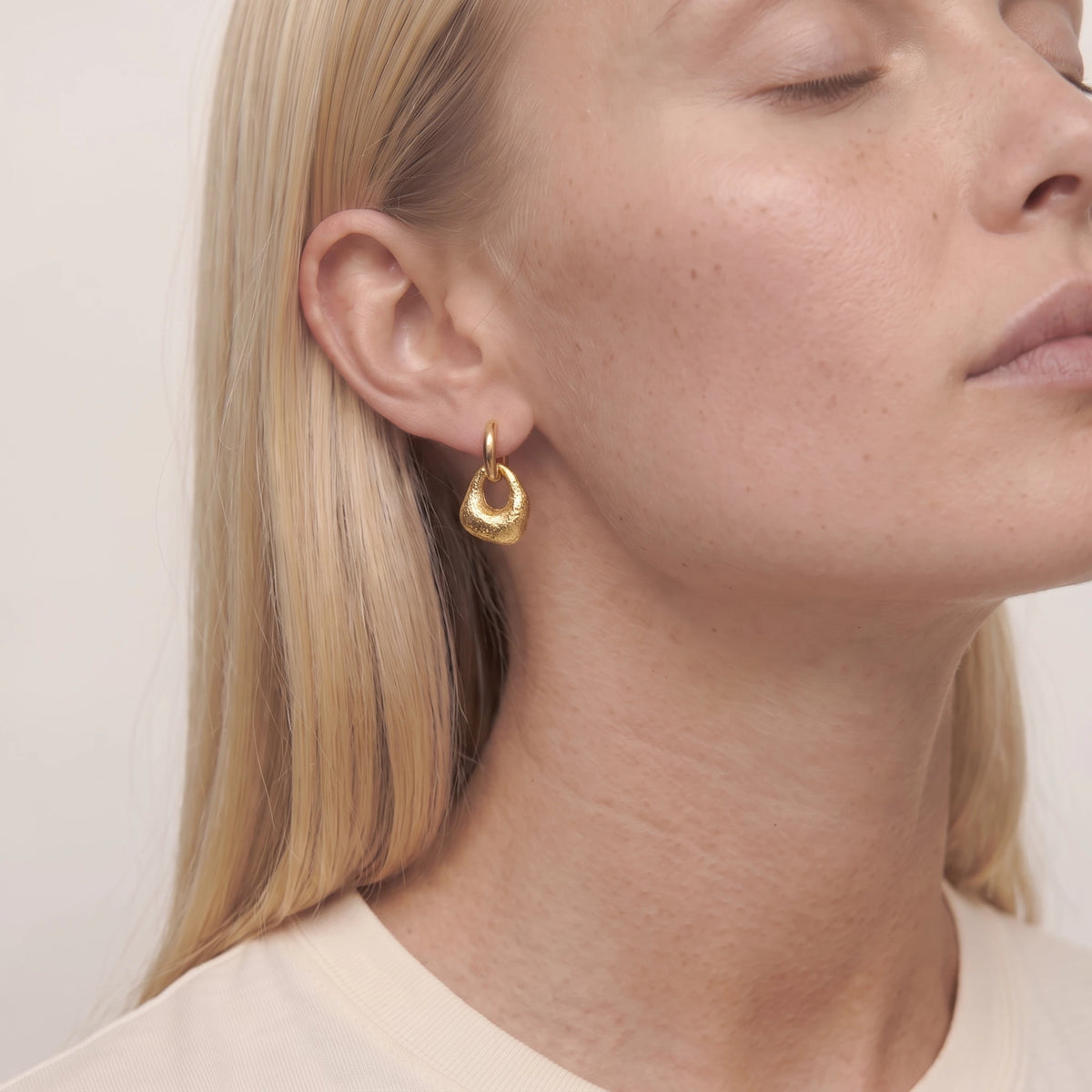 Close-up of a woman with her eyes closed wearing a gold Agnes molten drop huggie, in a cream top