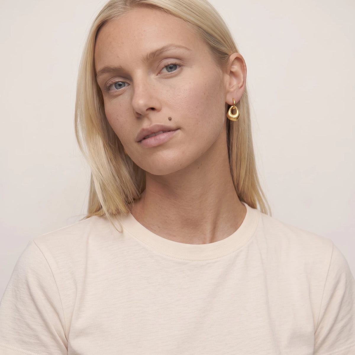 Woman with blonde hair and a beauty mark facing the camera, wearing a small gold Agnes drop huggie