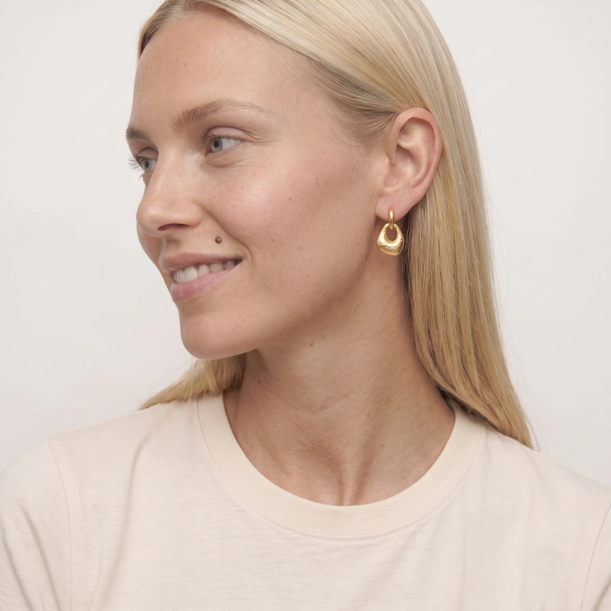 Woman with blonde hair smiling in side profile wearing a gold Agnes molten drop huggie, in a cream t-shirt